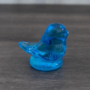 May include: A small, transparent, blue glass bird figurine. The bird is perched on a round base and has a smooth, glossy finish. The figurine is a decorative item, likely for display.