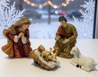 May include: A ceramic nativity set featuring a kneeling figure in a maroon and tan robe, a seated figure in a brown and orange robe, a baby in a tan manger, and a white lamb. The figures are painted in muted tones with detailed facial features.
