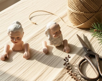 May include: Two porcelain doll parts sets featuring a female head with white hair and a male head with a white beard, each with two detached arms. Beside them are a ball of brown twine, a pair of metal scissors, and a small pine branch.