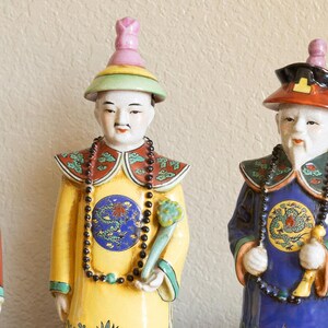 May include: Three porcelain figurines of people wearing traditional Chinese clothing. The figurines are painted in bright colors, including red, yellow, blue, and green. They are wearing elaborate hats and necklaces.