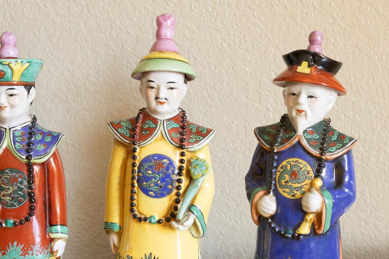 May include: Three porcelain figurines of people wearing traditional Chinese clothing. The figurines are painted in bright colors, including red, yellow, blue, and green. They are wearing elaborate hats and necklaces.