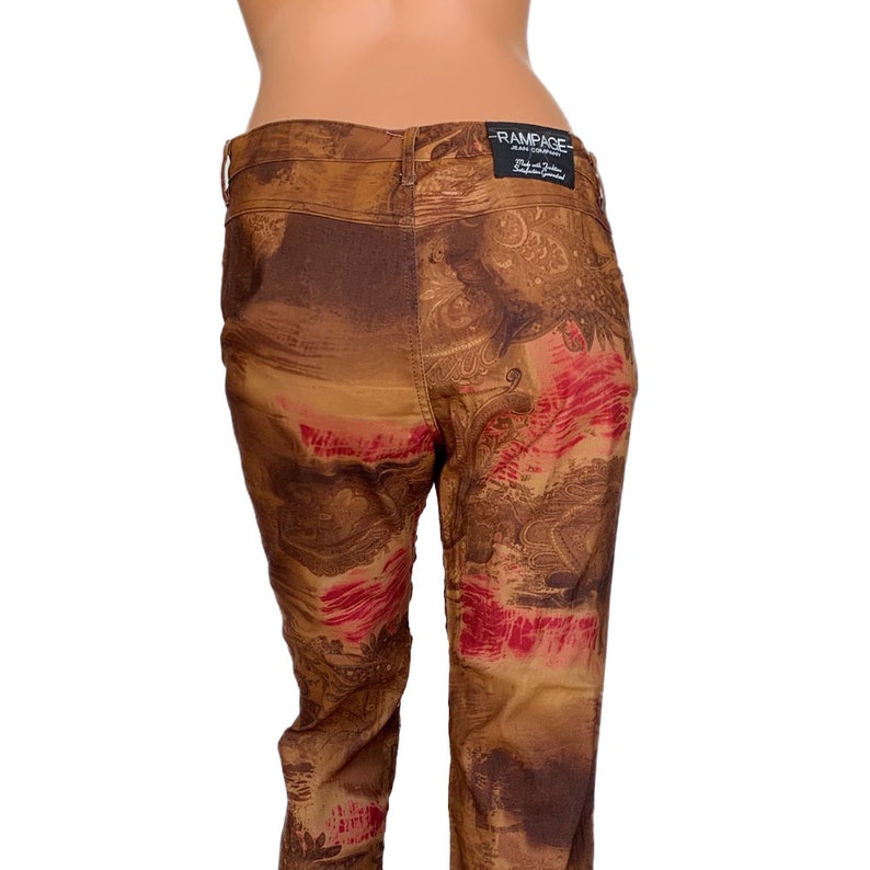May include: Brown jeans with a paisley print in shades of brown, red, and gold. The jeans have a back pocket with a "Rampage" label.