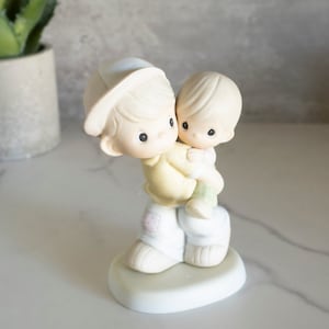 May include: A porcelain figurine of two boys, one carrying the other on his back. The boy in the front is wearing a baseball cap and has a yellow shirt and white pants. The boy on his back is wearing a white shirt and green pants. The figurine is on a white base.
