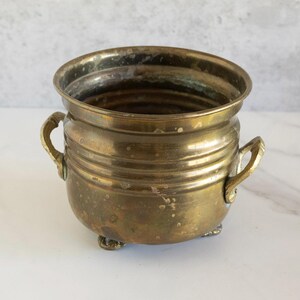 May include: A small, antique brass pot with two handles and three small feet. The pot has a rounded shape with horizontal ridges around the body. The brass has a tarnished, aged appearance, suggesting it is vintage.