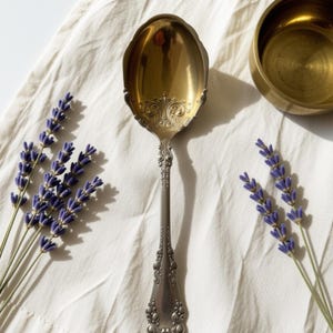 May include: A silver-toned metal spoon with a gold-colored bowl featuring intricate embossed scrollwork at the base of the handle. The spoon is positioned vertically between two sprigs of purple lavender flowers and a small gold-toned metal bowl.