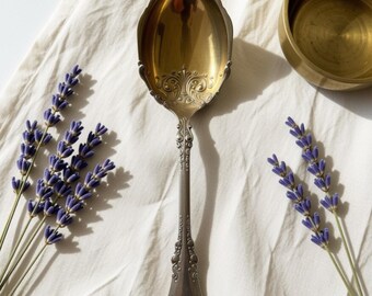 May include: A silver-toned metal spoon with a gold-colored bowl featuring intricate embossed scrollwork at the base of the handle. The spoon is positioned vertically between two sprigs of purple lavender flowers and a small gold-toned metal bowl.