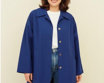 May include: A long, royal blue coat featuring a classic collar, long sleeves, and a front button closure. The coat is worn over a plain white crew neck t-shirt and blue denim jeans, creating a simple, layered look with visible side pockets.