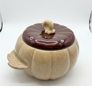 May include: A ceramic pumpkin-shaped container with a lid. The container is tan with a brown lid and a beige handle. A leaf-shaped detail is on the side. The container is likely for food storage or decoration.