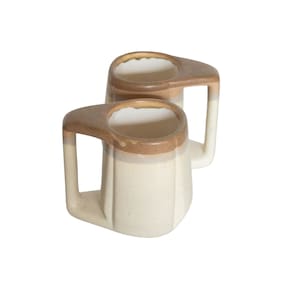May include: Two ceramic mugs with brown and beige glaze. The mugs have a wide, rounded base and a handle that curves up and over the rim.
