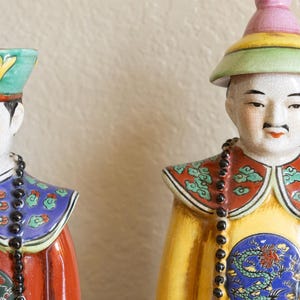 May include: Two porcelain figurines of people wearing traditional Chinese clothing and hats. The figurines are painted with intricate details and bright colors, including red, yellow, blue, and green. Both figurines are holding black beaded necklaces.