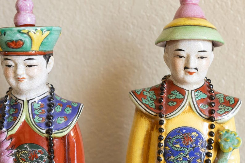 May include: Two porcelain figurines of people wearing traditional Chinese clothing and hats. The figurines are painted with intricate details and bright colors, including red, yellow, blue, and green. Both figurines are holding black beaded necklaces.