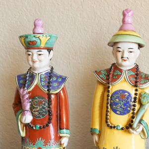 May include: Two porcelain figurines of people wearing traditional Chinese clothing. The figurines are painted in bright colors, including red, yellow, blue, and green. They are holding objects in their hands, and their faces are painted with expressions of serenity.