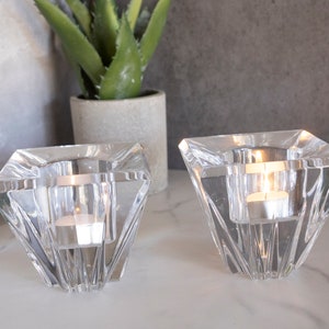 May include: Two clear glass candle holders with a geometric diamond shape. The holders are empty and sitting on a white surface.