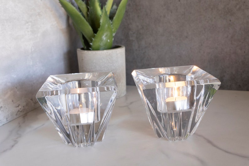 May include: Two clear glass candle holders with a geometric diamond shape. The holders are empty and sitting on a white surface.