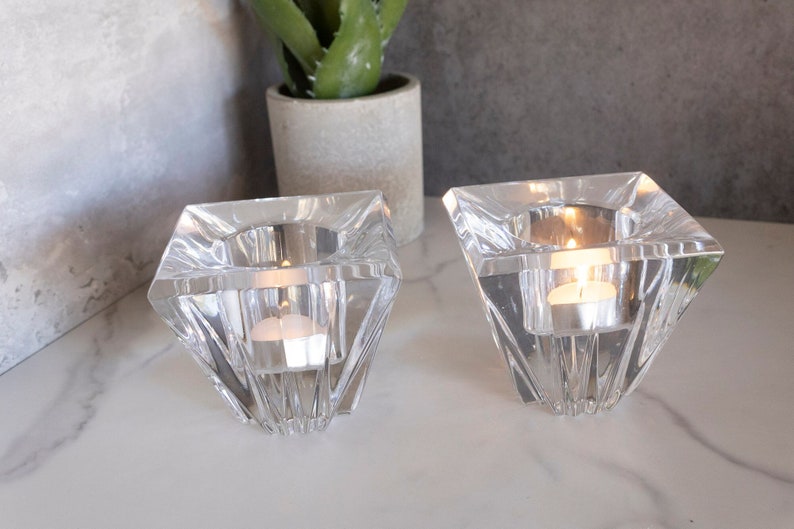 May include: Two clear glass candle holders with a geometric design. Each holder has a square base and a pointed top, creating a diamond shape. The holders are filled with tealight candles.