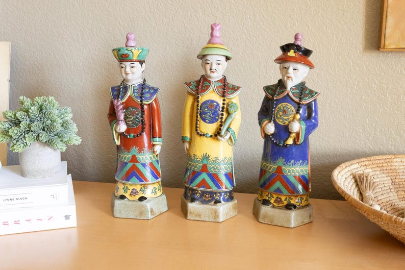 May include: Three porcelain figurines of people wearing traditional Chinese clothing. The figurines are painted in bright colors, including red, yellow, blue, and green. They are standing on a wooden surface.