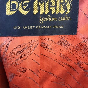 Puede incluir: Una etiqueta negra con letras doradas que dice "De Mark's Fashion Center" y "6101 West Cermak Road".