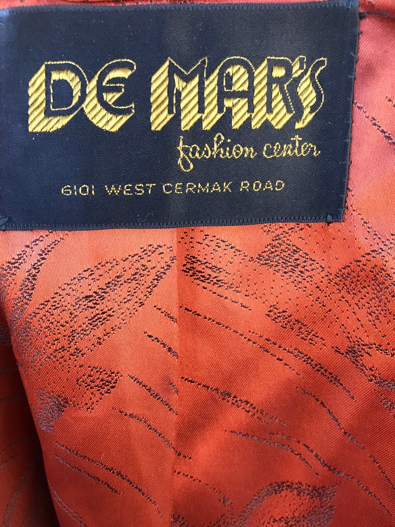Puede incluir: Una etiqueta negra con letras doradas que dice "De Mark's Fashion Center" y "6101 West Cermak Road".