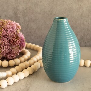 May include: A teal blue ceramic vase with a ribbed texture. The vase is sitting on a table with a wooden bead garland and a white tassel.