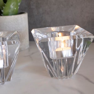 May include: Two clear glass candle holders with a geometric design. The holders are shaped like a pyramid with a square base. Each holder has a small, round opening at the top for a tea light candle.