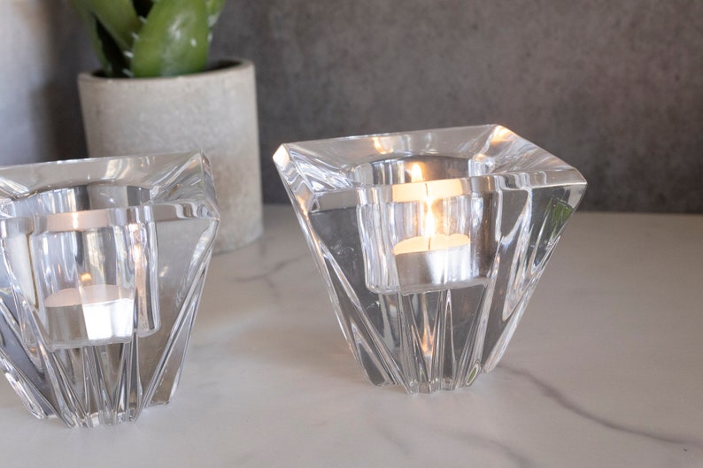 May include: Two clear glass candle holders with a geometric design. The holders are shaped like a pyramid with a square base. Each holder has a small, round opening at the top for a tea light candle.