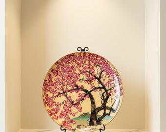 May include: A round decorative plate featuring a painted scene of a tree with vibrant pink blossoms against a pale yellow background. The tree has a dark brown trunk and branches, with a small green landscape at the base. The plate has a gold rim.