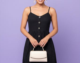 May include: A black midi dress with thin spaghetti straps, a V-neckline, and a vertical row of buttons down the center front. The dress features a fitted bodice and a flared skirt. A small, structured, off-white handbag with a top handle is held.
