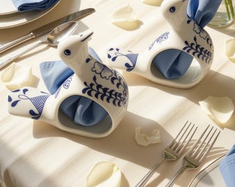 May include: Two white ceramic napkin rings shaped like birds, each featuring intricate blue floral and foliate patterns. The hollow bodies of the bird-shaped holders contain folded light blue cloth napkins, creating a decorative table setting.
