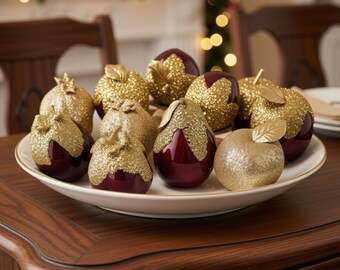 May include: A collection of decorative fruit ornaments arranged on a white ceramic plate. The pieces include deep burgundy apples and pears partially covered in textured gold glitter, alongside solid gold glittered apples with gold leaves.