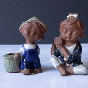 May include: Two ceramic figurines of children. The boy is wearing a blue and white outfit and a straw hat. He is holding a basket. The girl is wearing a blue and white kimono and has a white bow in her hair. She is holding a small drum.