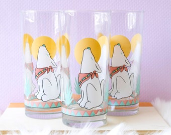 May include: Set of three clear glass tumblers with a design of a white wolf howling at a yellow moon. The wolf is wearing a red, blue, and green bandana. The glasses have a turquoise and pink border.