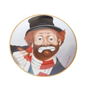 May include: A porcelain plate featuring a colorful portrait of a clown with a sad expression. The clown is wearing a black top hat, a red and white scarf, and holding a cigar. The plate has a gold rim and the artist's signature is visible in the bottom right corner.