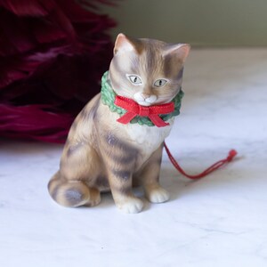 May include: A ceramic cat figurine wearing a green wreath with a red bow. The cat is sitting on a white marble surface.