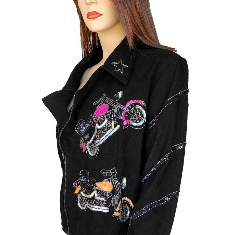 K&ouml;nnte beinhalten: Schwarze Jacke mit Pailletten-Motorrad-Stickerei. Die Jacke hat einen Rei&szlig;verschluss und eine sternf&ouml;rmige Verzierung am Kragen.