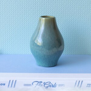 May include: A small, teal-colored ceramic vase with a rounded, bulbous shape. The vase has a subtle, iridescent glaze that shimmers in the light.