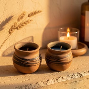 May include: Two small, round, terracotta-colored clay pots containing dark wax with a single centered wick in each. The pots feature horizontal, dark brown, swirling, painted brushstroke patterns around their midsections.