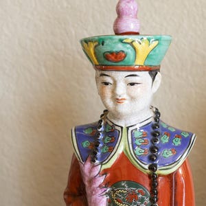 May include: A porcelain figurine of a person wearing a colorful, ornate hat and robe. The figurine has a beaded necklace and a painted floral design on the robe.