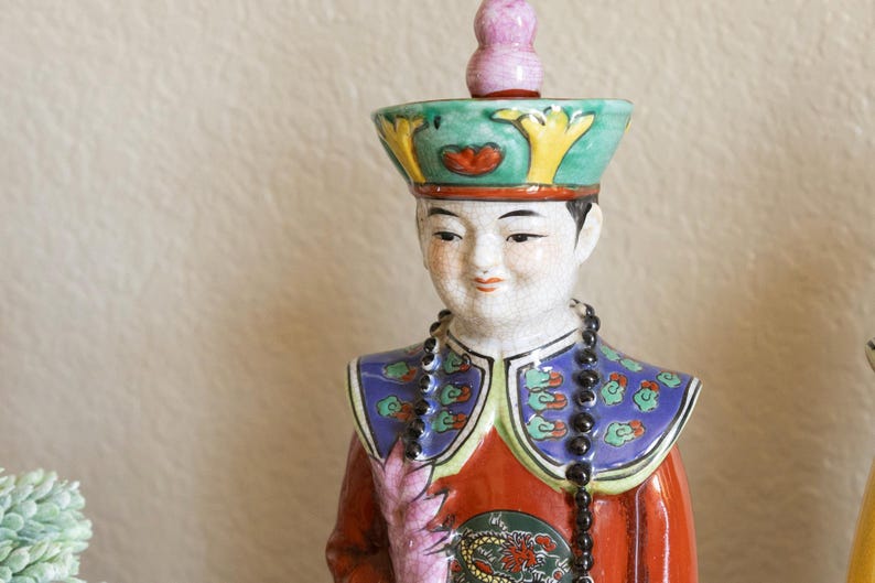 May include: A porcelain figurine of a person wearing a colorful, ornate hat and robe. The figurine has a beaded necklace and a painted floral design on the robe.