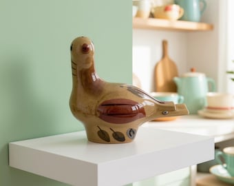May include: A ceramic bird figurine featuring a glossy tan glaze with dark brown painted details on the wings and body. The bird has a rounded head, a small beak, and stylized leaf patterns painted along the lower base of the figure.