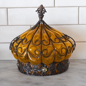 May include: An ornate, amber-colored glass jar with a decorative metal filigree cage and base. The jar has a domed shape with a detailed finial on top. The base features floral and jewel accents, creating a vintage aesthetic.