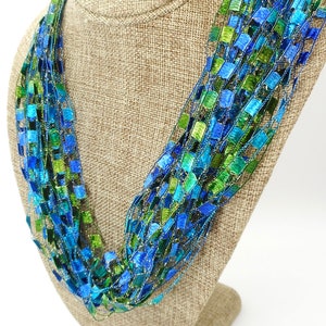May include: A multi-strand ribbon necklace featuring a woven design of metallic blue, turquoise, and lime green rectangular segments. The necklace is constructed from thin, shimmering threads that hold the colorful ribbon pieces in a loose, layered arrangement.