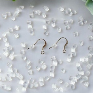 May include: Two gold earring hooks with a clear silicone stopper on a white surface. There are many clear silicone stoppers scattered around the hooks.