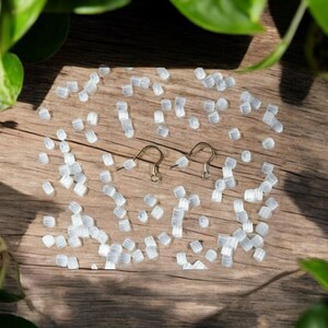 May include: A close-up of a wooden surface with a scattering of small, white, plastic beads. Two silver earring hooks are placed in the centre of the image.