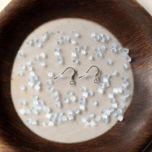 May include: A close-up of a white ceramic plate with a brown wooden rim. The plate is filled with small, clear, plastic earring backs. Two silver earring hooks are also on the plate.