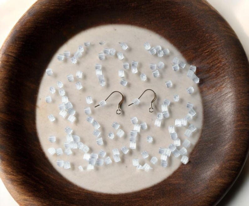 May include: A close-up of a white ceramic plate with a brown wooden rim. The plate is filled with small, clear, plastic earring backs. Two silver earring hooks are also on the plate.