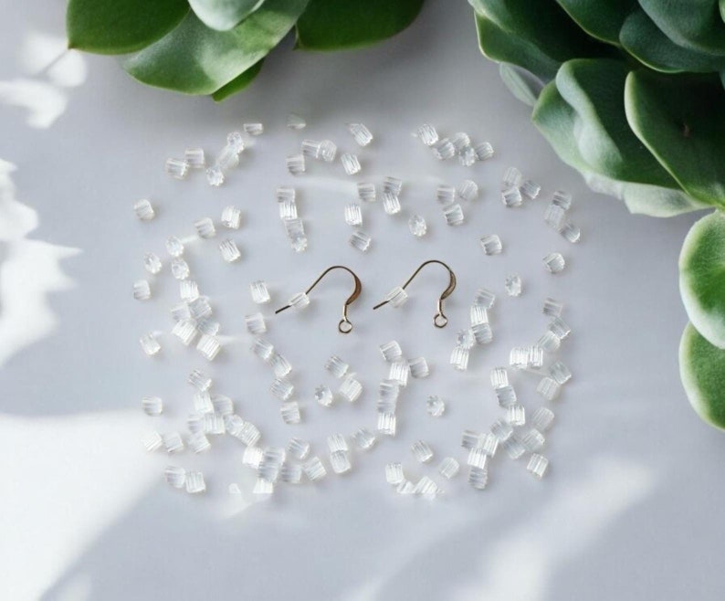May include: Two gold earring hooks with a clear silicone stopper on a white surface. There are many clear silicone stoppers scattered around the hooks.