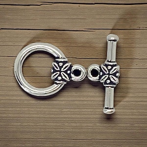 May include: Silver-toned toggle clasp with a circular ring and a bar, both adorned with floral-like designs. The clasp is set against a wooden background, showcasing its intricate details and metallic sheen.