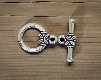May include: Silver toggle clasp featuring a circular ring and a bar, both decorated with floral patterns. The clasp is set against a wooden background, highlighting its intricate design and metallic finish.