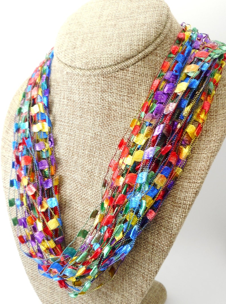 May include: A multi-strand necklace made of colourful, iridescent ribbon. The necklace is displayed on a brown fabric mannequin.