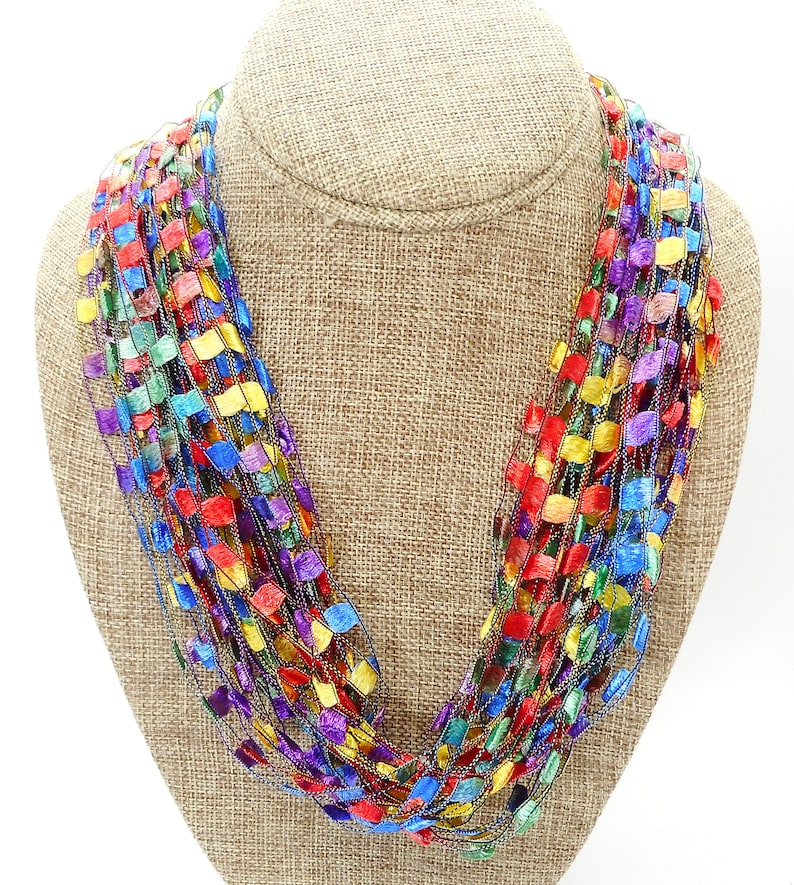 May include: A multi-coloured crocheted necklace with a metallic sheen. The necklace is made of a variety of colours, including red, orange, yellow, green, blue, and purple.
